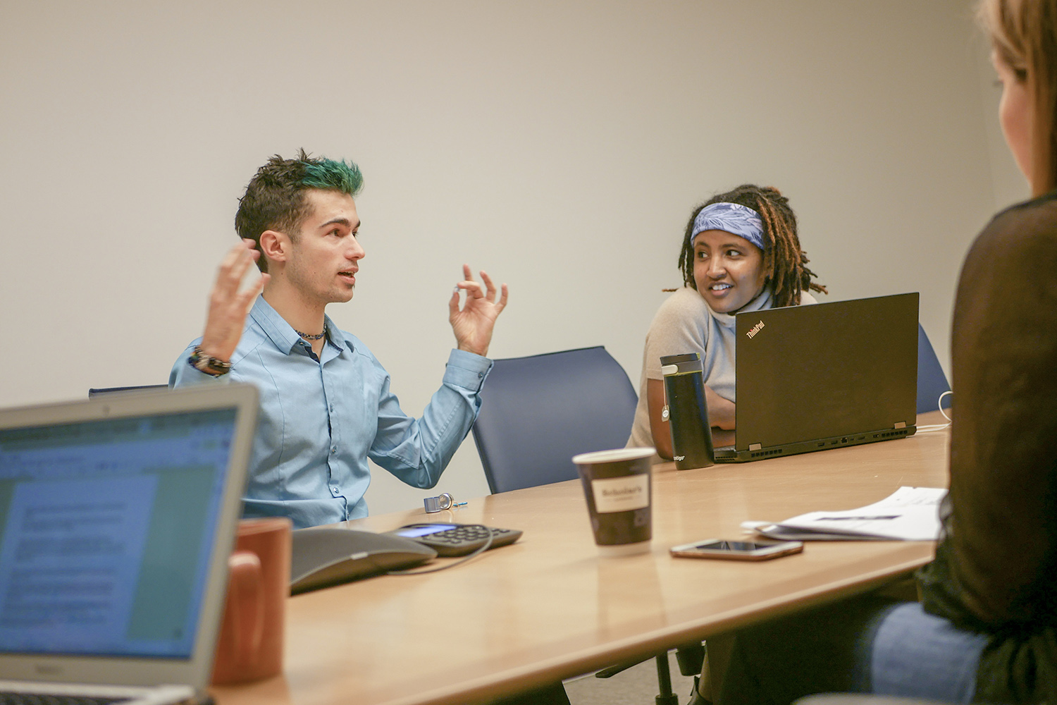 A person gestures while speaking to two colleagues at a meeting table with laptops and coffee cups.