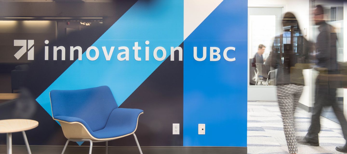 A wall painted with blue colours and the innovation UBC logo, with an empty blue chair and two people walking