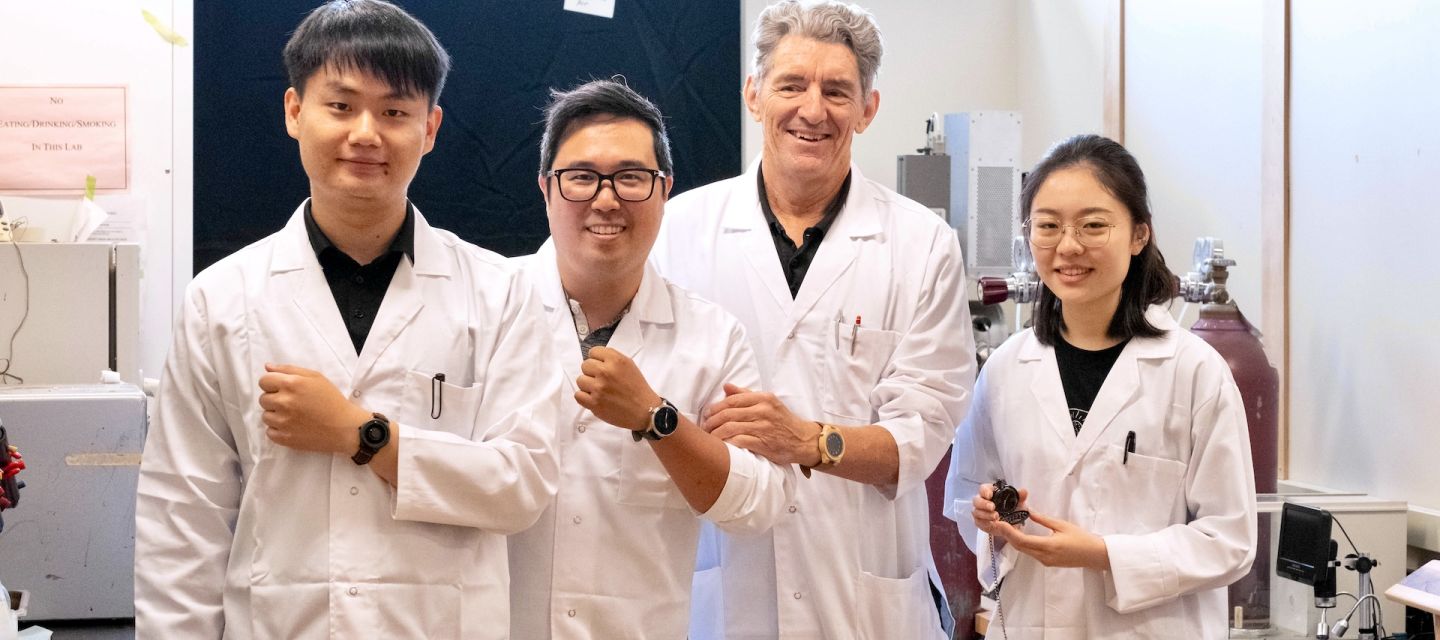 Three males and one female stand side-by-side in labcoats, in a lab, each with their left arm bend and showing off a watch.