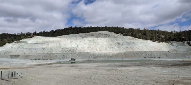 Carbon removal research site at a legacy mine in western US: white sandy hill with coniferous trees and cloudy sky are behind.