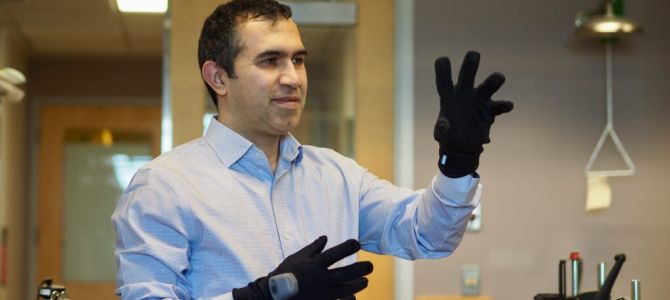 a man in a blue, collared dress shirt holds up his hands, wearing thick black gloves that have small metallic-looking sensors on their back. He is looking to the right at his raised hands. 