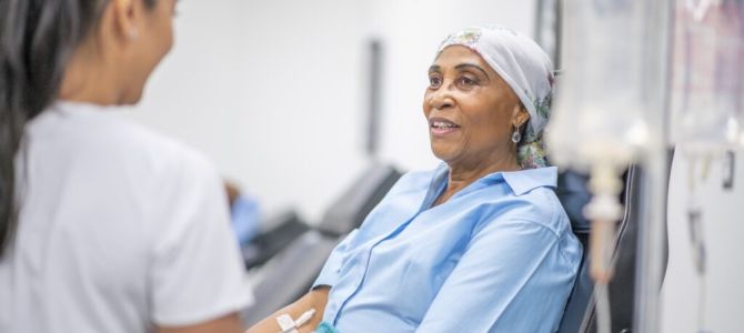 Woman in blue hospital gown and white medical cap lies in a bed looking up at a woman in a white hospital uniform, who has long dark hair that is up in a ponytail.