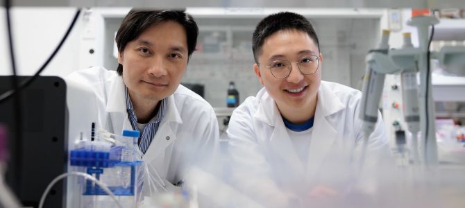 Two Asian males in lab coats stand side-by-side looking toward the camera, with lab equipment including test tubes and machines. 