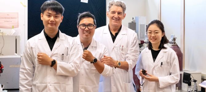 Three males and one female stand side-by-side in labcoats, in a lab, each with their left arm bend and showing off a watch.