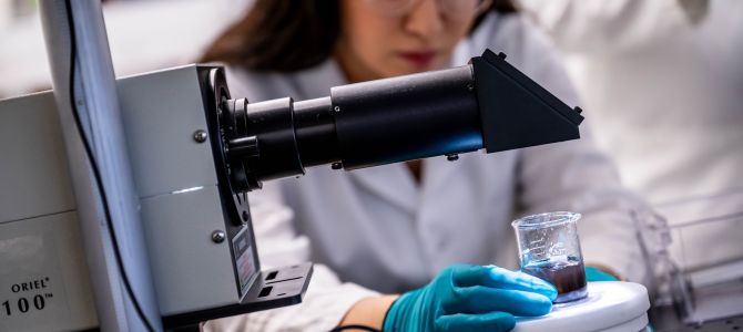 woman in white labcoat and blue medical gloves looks intently at a small beaker filled with a clear blue-tinted liquid.